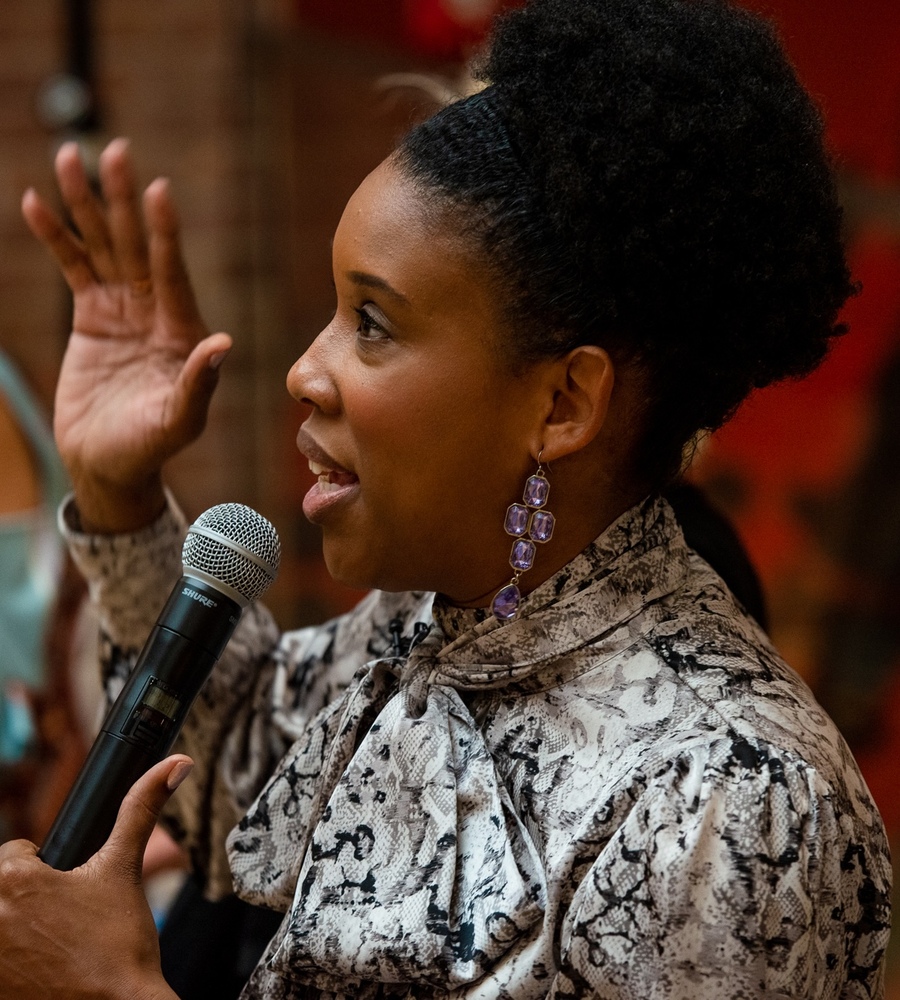 Side profile of comedian Joyelle Nicole Johnson speaking into a microphone with her hand raised at the Funny Ladies: Serious Business event.