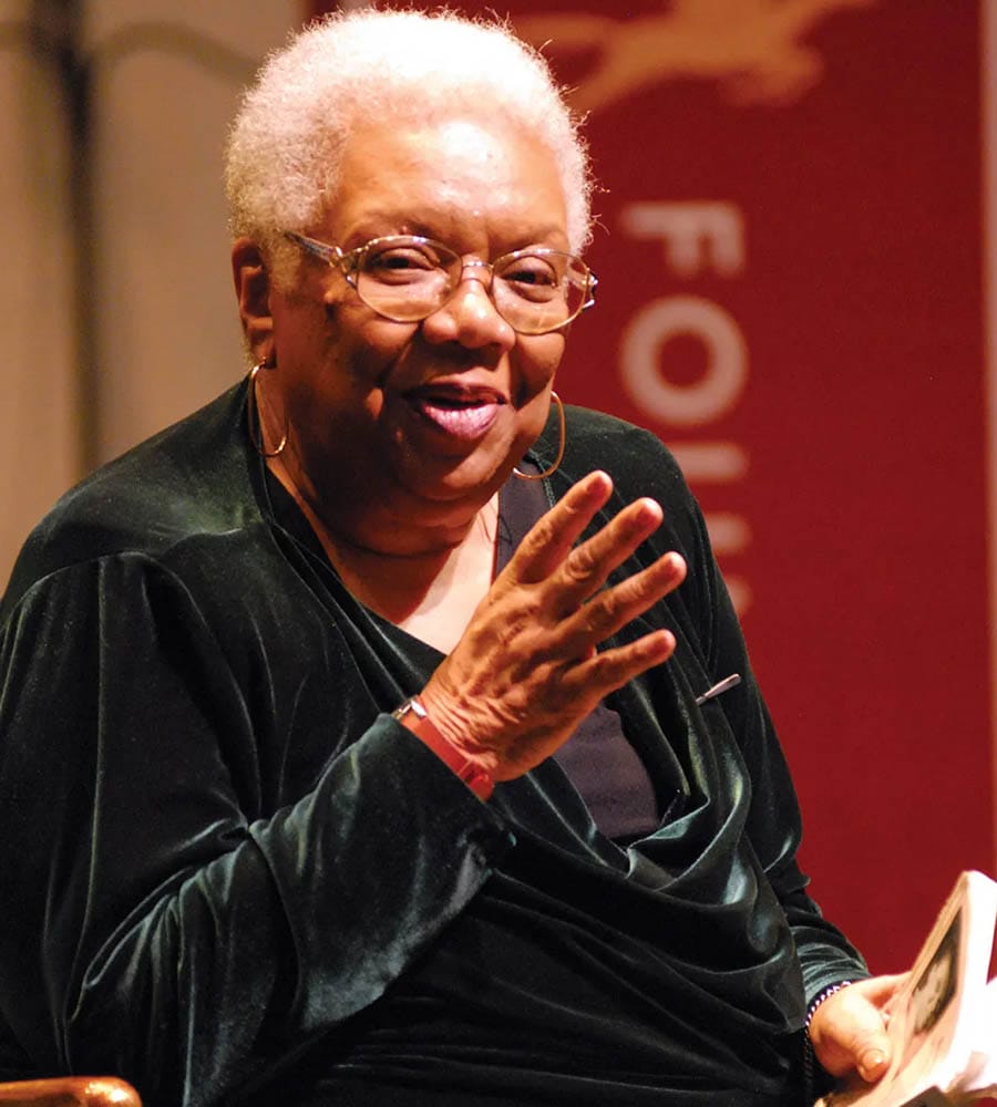 Lucille Clifton, an African American poet and educator, gestures while speaking at the Geena Davis Institute event, wearing glasses and a black velvet top, radiating wisdom and engagement in a warm-lit room.