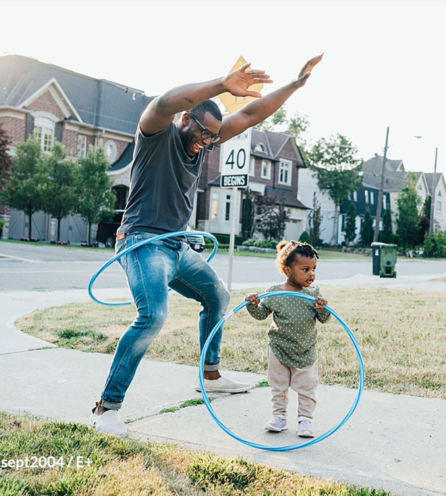 A man and a toddler engaging in a hula hoop activity on a residential street, both smiling and having fun in a sunny outdoor setting.