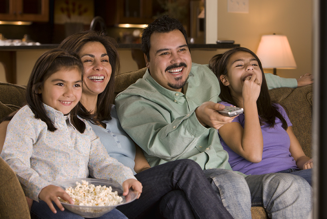 A happy family enjoys movie night at home, with a mother and father seated on the couch with their two daughters. The father holds a remote control, while the youngest daughter holds a bowl of popcorn, and they all share a joyful moment watching television.