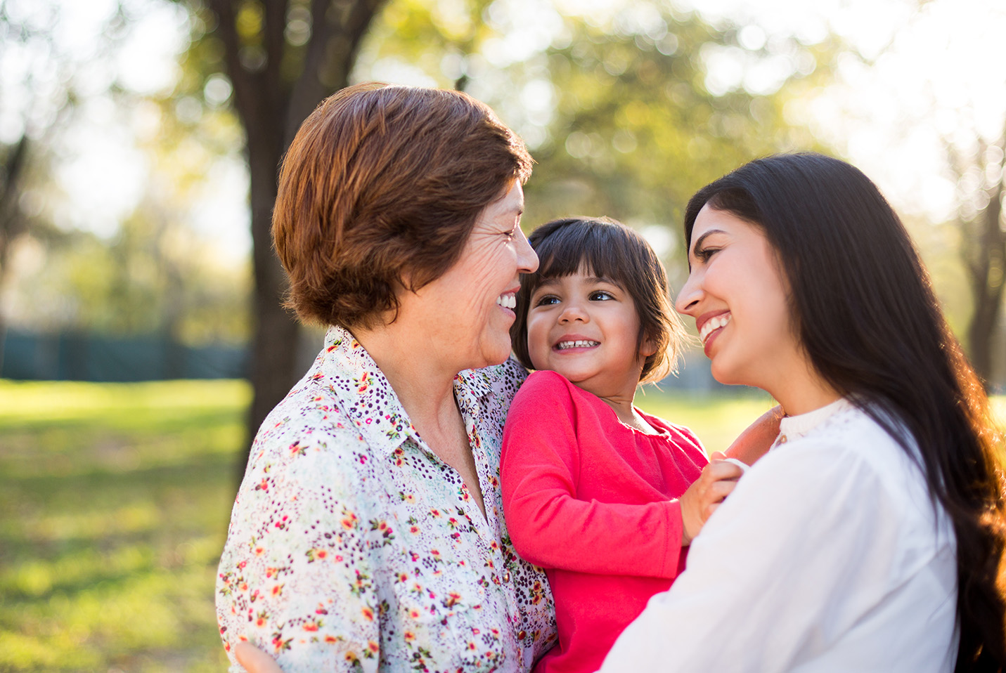 Three generations of a family share a joyous moment outdoors; an elderly woman with a floral shirt smiles at a young girl in red, who is held by a smiling young woman in white. The backdrop is a sunlit park with trees, suggesting a warm, familial atmosphere.