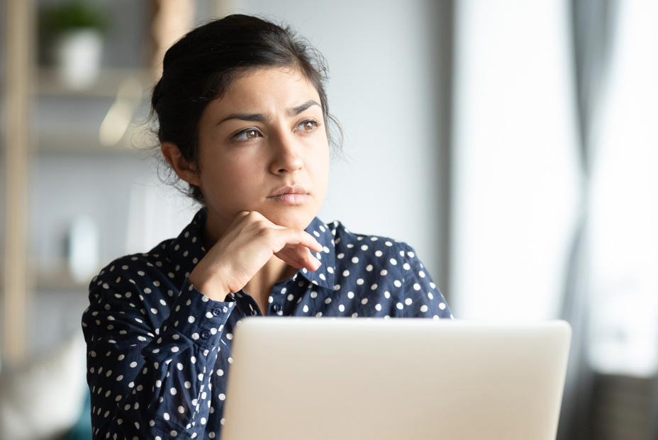 A pensive young woman in a polka dot blouse holds a pen near her mouth, looking away with a contemplative expression while working on her laptop in a bright, modern office setting.