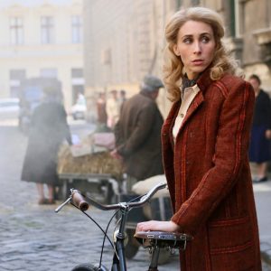 A woman in a vintage red coat stands next to a bicycle on a cobblestone street, looking over her shoulder with a serious expression. The background shows a bustling historical setting with people and carts.