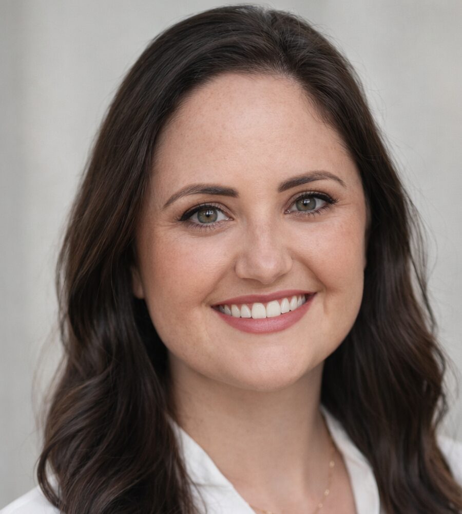 A professional headshot of Jennie de la Sotta, a woman with wavy dark brown hair and a warm smile, wearing a white blouse and gold necklace against a neutral gray background.