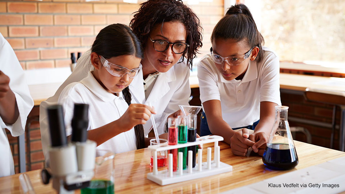 A science teacher assists two young students with a chemistry experiment in a classroom, with test tubes filled with colorful liquids on the table and a microscope on the side. Safety glasses are worn by all, indicating a hands-on educational science activity.