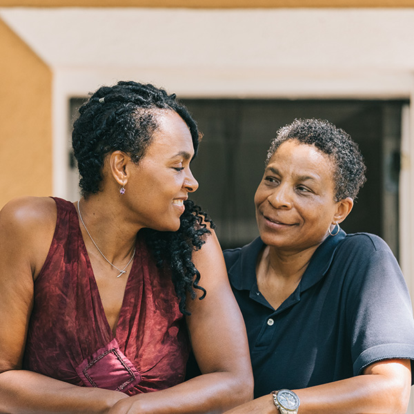 Two women in their 50s sit close together outdoors, smiling and gazing warmly at each other. One woman is wearing a sleeveless burgundy top, while the other wears a navy blue polo shirt.