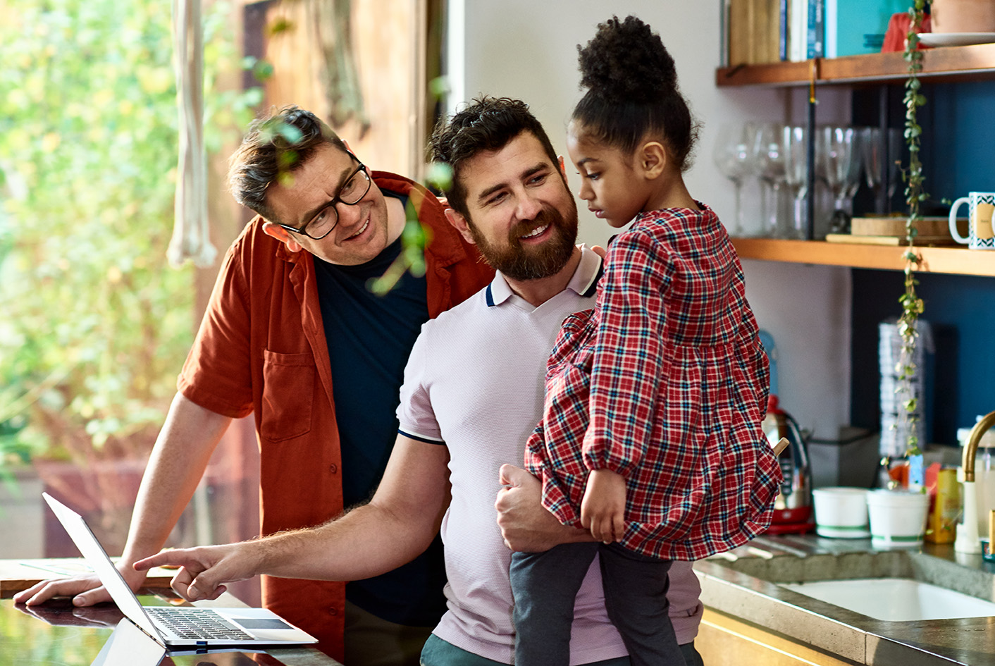 Three people stand in a home kitchen, focused on a laptop screen. A man with a beard holds a young girl in his arms, while another man points at something on the screen. They are all engaged and appear to be in a positive, comfortable home environment.