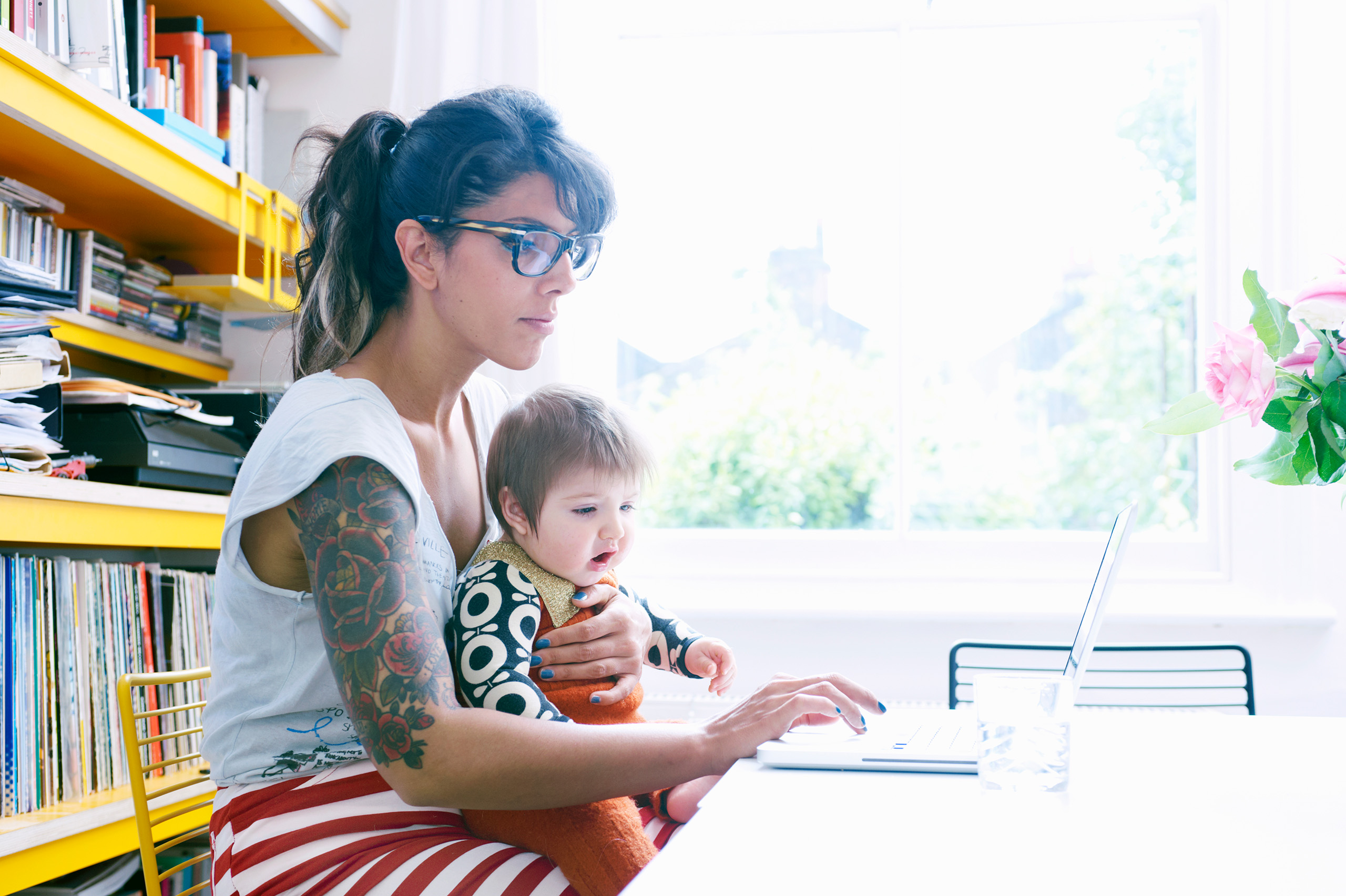 Motherhood exemplified by a woman with a child on her lap working at a laptop beside a window.