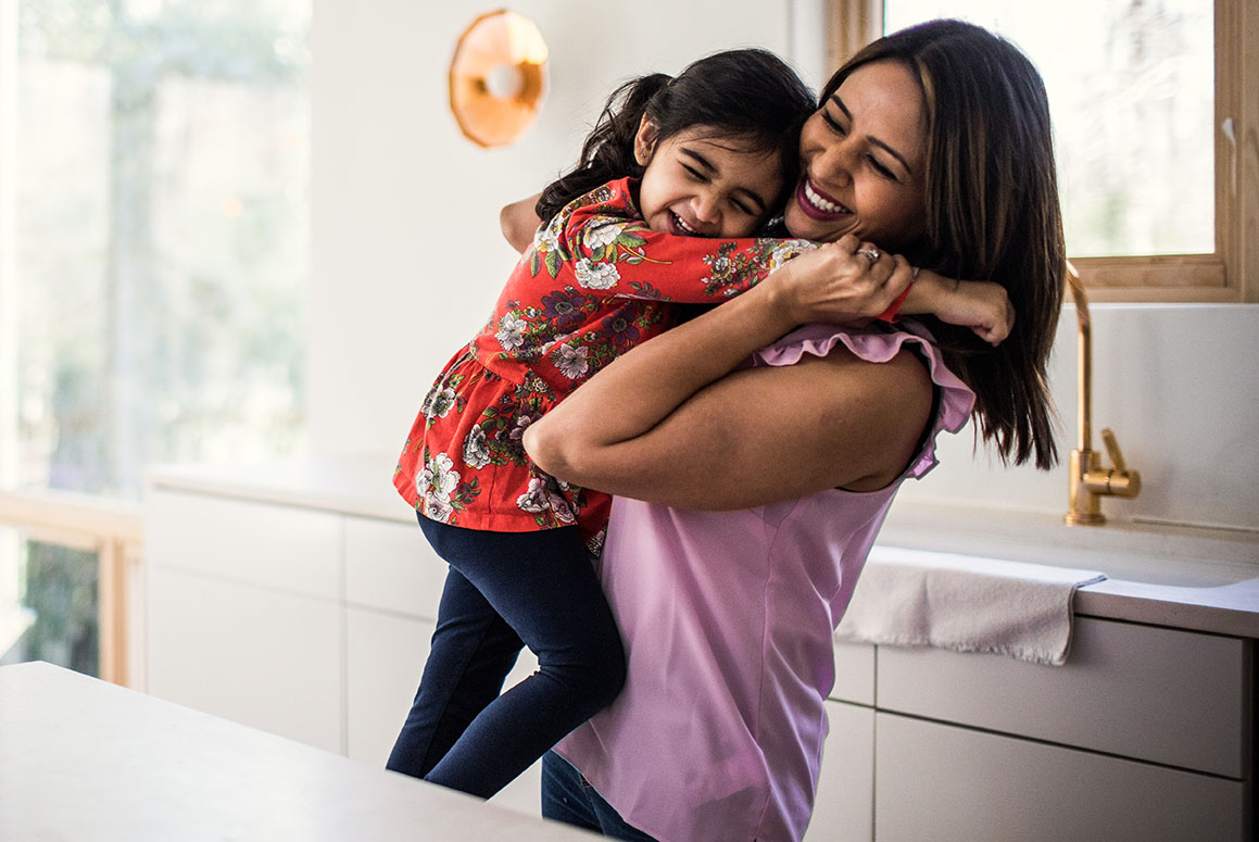 Alt text: A joyful moment as a mother in a lavender top hugs her daughter, who is giggling and dressed in a red floral top and blue jeans, in a well-lit kitchen setting with modern decor.