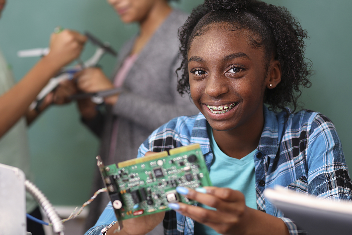 A young student with braces on her teeth smiles at the camera, holding a green circuit board as she participates in a robotics or electronics class, with another student working in the background. The focus is on hands-on STEM education and the joy of learning.