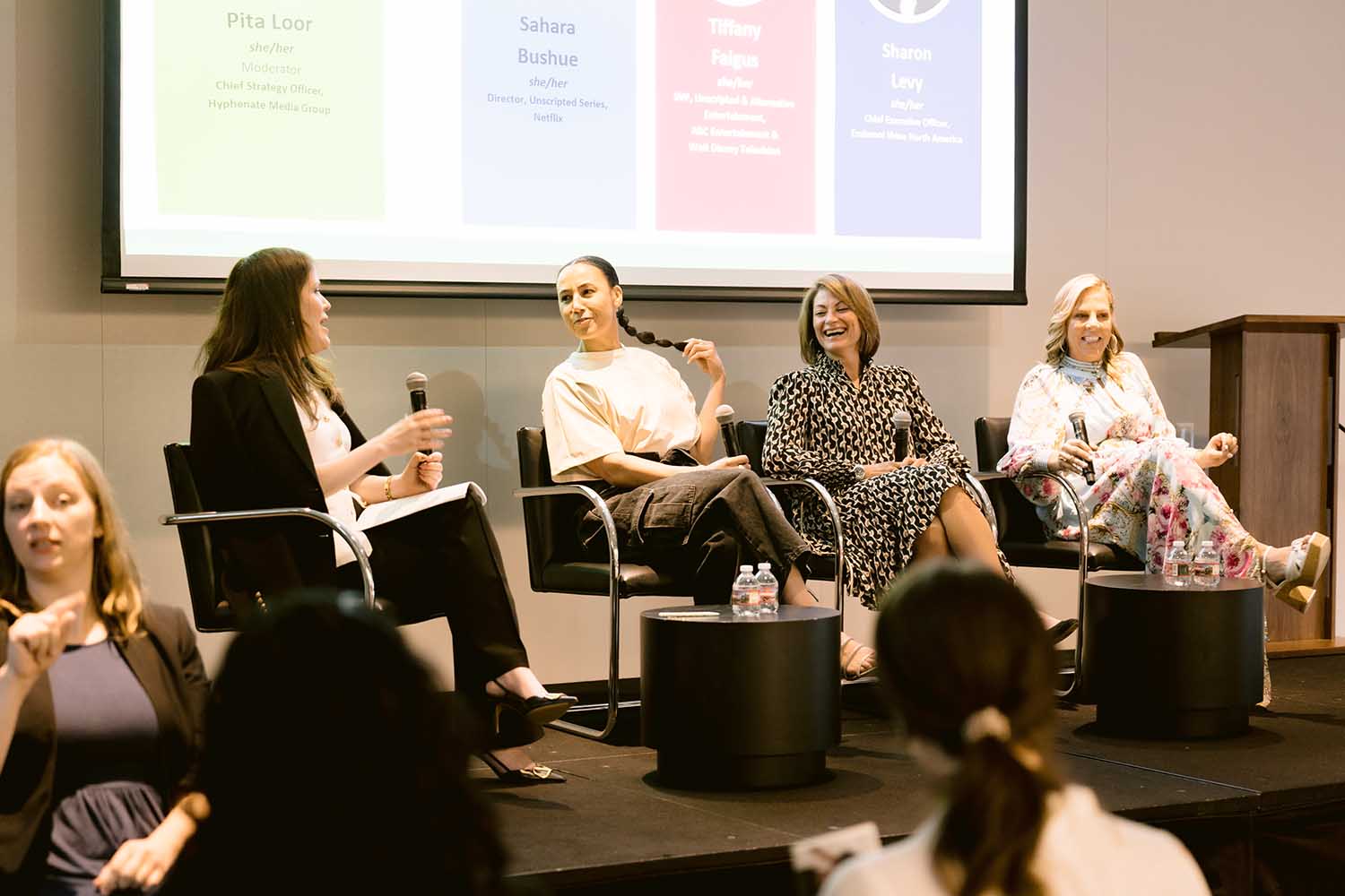 Four women sit on a stage during a panel discussion. They are smiling and holding microphones. Behind them is a screen with names and titles. The audience is seated and engaged.