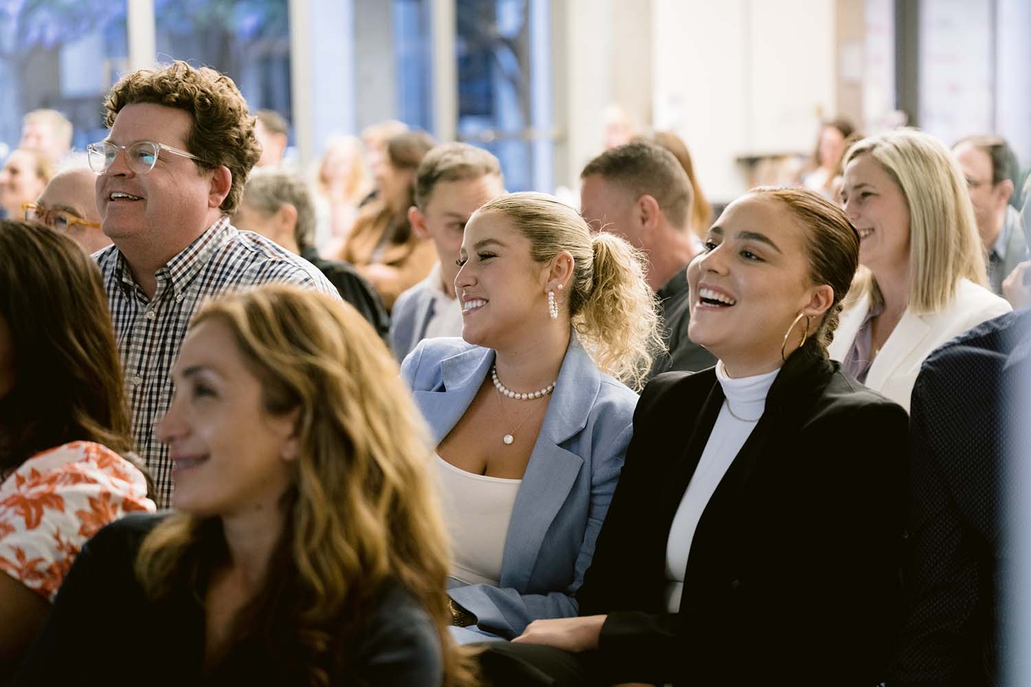 A diverse group of attendees seated and smiling during an event. The audience appears engaged and entertained. The setting is a well-lit indoor venue, with several rows of people visible in the background.