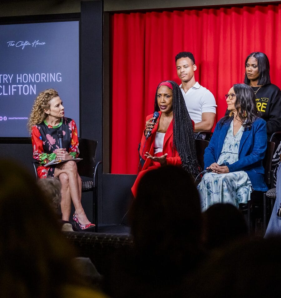 Panel discussion at The Geena Davis Institute event with a diverse group of individuals on stage, including a woman speaking into a microphone, against a backdrop of red curtains and a screen displaying 'The Clifton House.