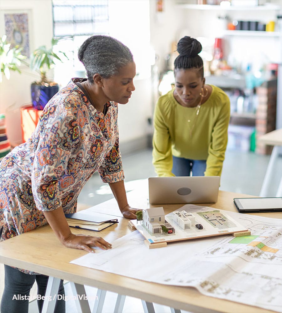 Two women in a bright workspace review architectural plans and a small model house on a table, with a laptop nearby, symbolizing collaboration in design or planning.