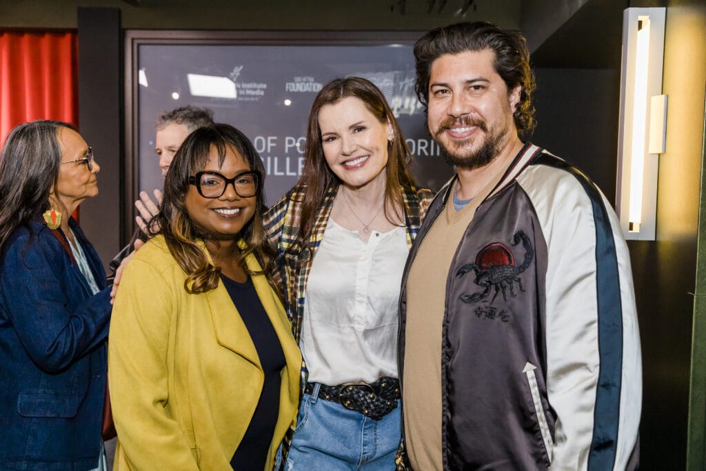Wendy Calhoun, Geena Davis and Narumi Inatsugu posing together at an event, with a woman in a yellow blazer and glasses in the foreground, flanked by another woman in a plaid jacket and jeans, and a man in a bomber jacket with an embroidered scorpion, all smiling at the camera with event signage in the background