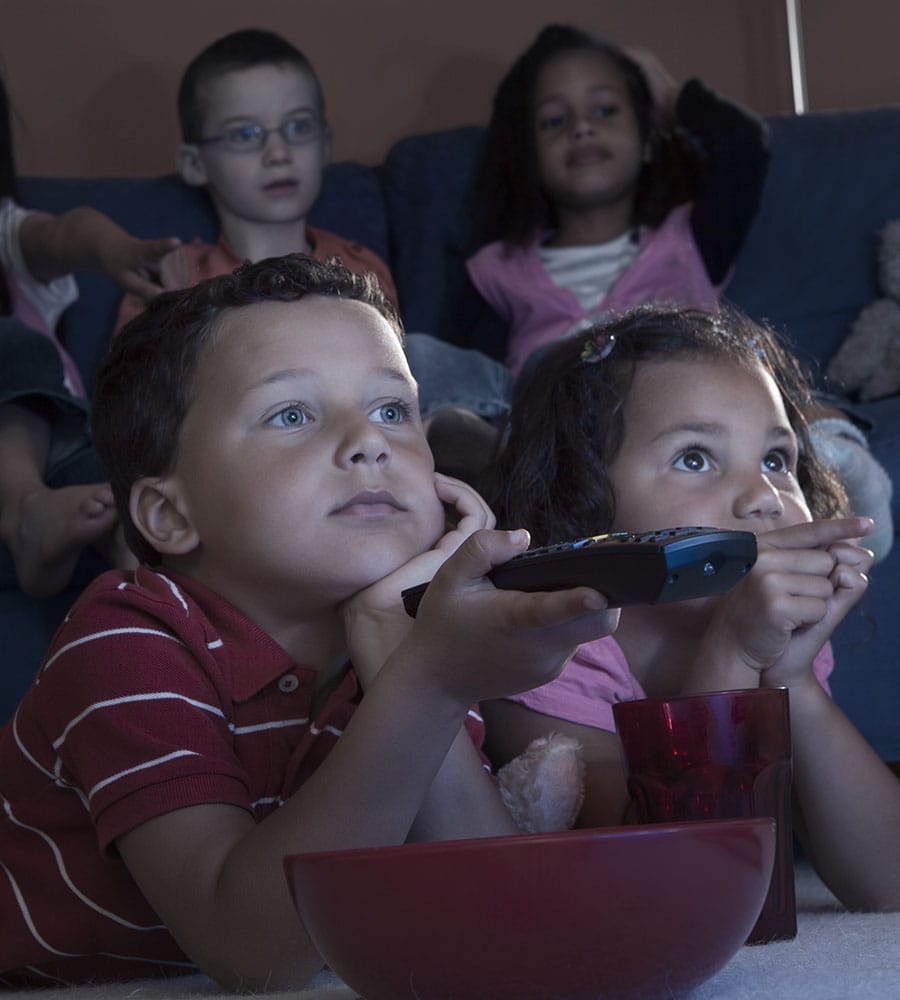 A group of children intently watching television in a dimly lit room, two of them holding a remote, with a bowl of snacks in the foreground. Their expressions are a mix of excitement and concentration, reflecting the engaging content on the screen.