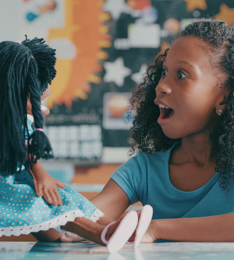 A young girl with curly hair, wearing a blue shirt, appears excitedly speaking to a doll seated across from her at a small table. They are in a classroom setting with a star-shaped decoration in the background.