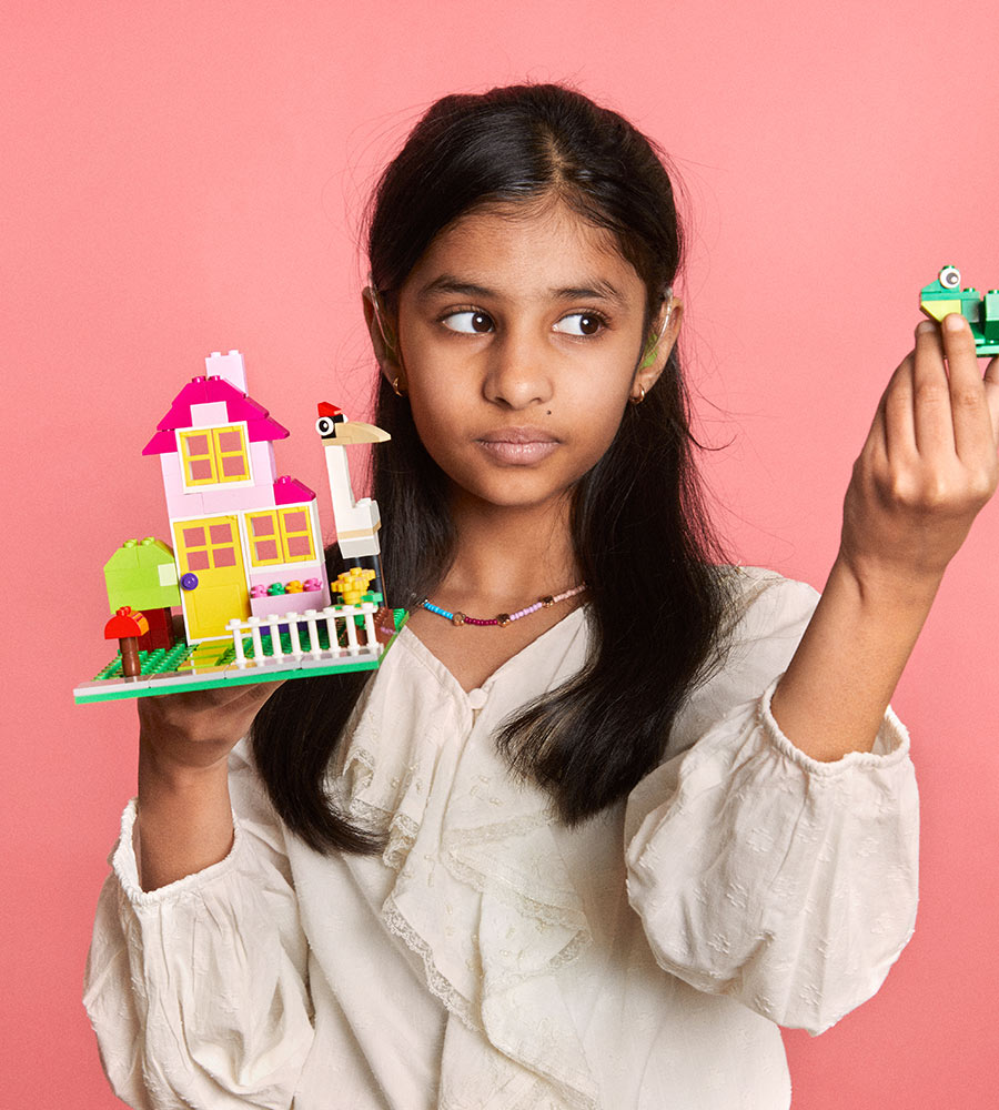 A young girl holding a colorful LEGO house on one hand and a green LEGO frog on the other, wearing a casual white blouse with a beaded necklace, against a pink background.
