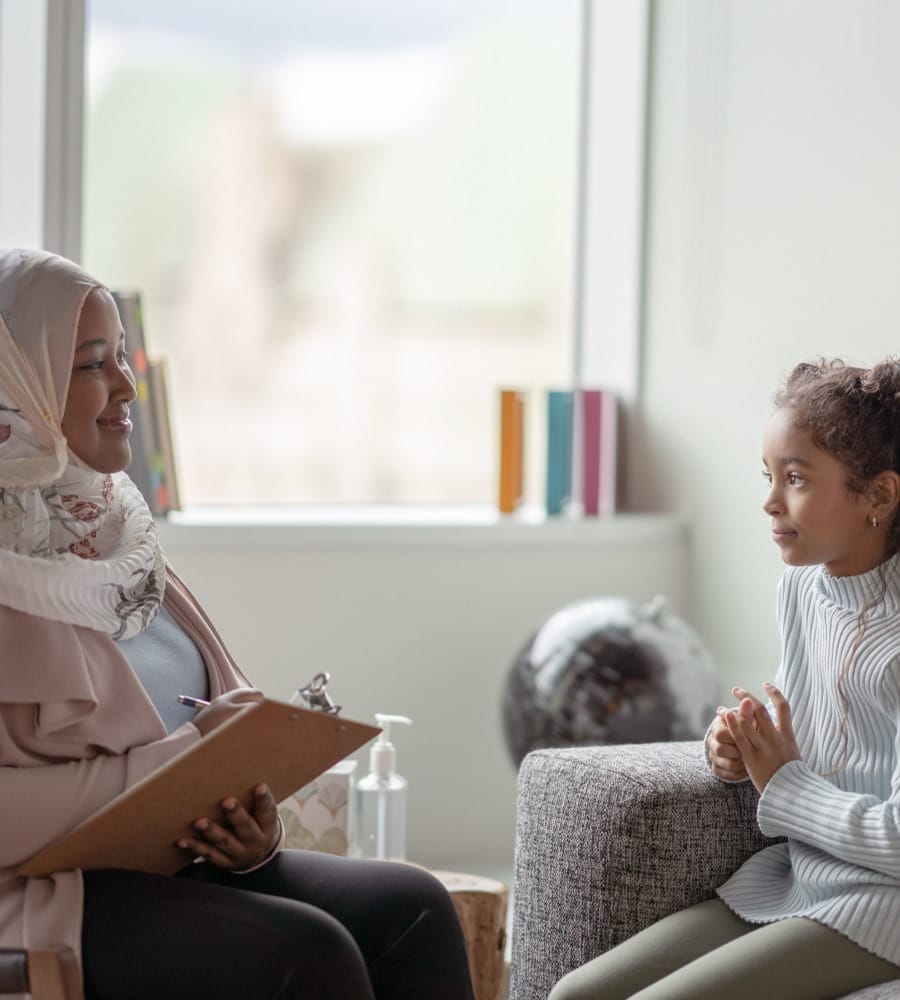 A Muslim therapist sits with her patient. The young girl is seated on a sofa while the therapist sits across from her on a chair with a clipboard in her hands.