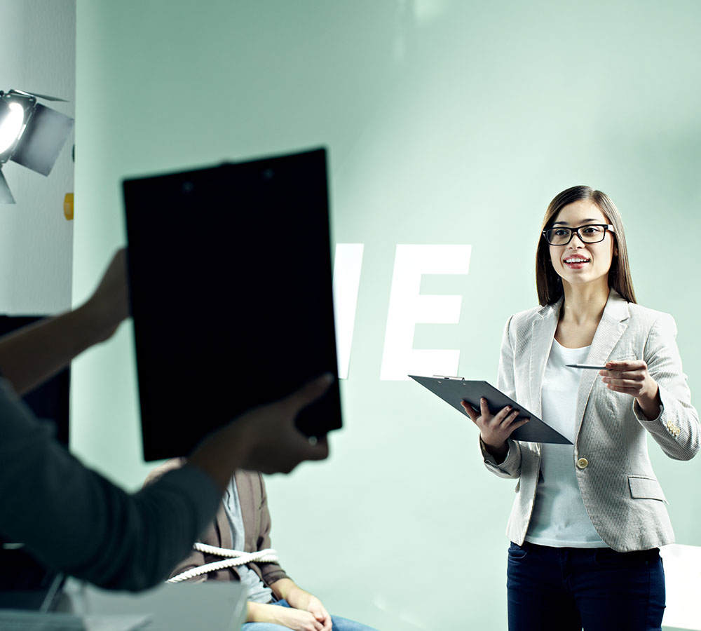 A woman in a light-colored blazer and glasses holds a clipboard and pen, speaking and gesturing towards someone out of frame in a bright, well-lit room. Another person in the foreground holds a black clipboard, partially obscuring the background.