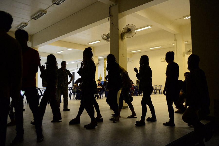 Silhouettes of women and others stand in line inside a dimly lit institutional room with empty chairs in the background. The scene creates a somber and reflective atmosphere, with a focus on the women’s shapes and movement.