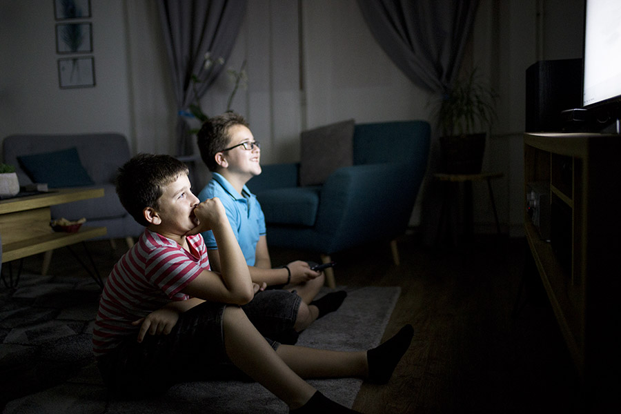 Two young boys sit on the floor in a dimly lit living room, watching TV intently. One boy leans forward with a big smile, while the other, wearing glasses, holds a remote and watches with equal excitement.