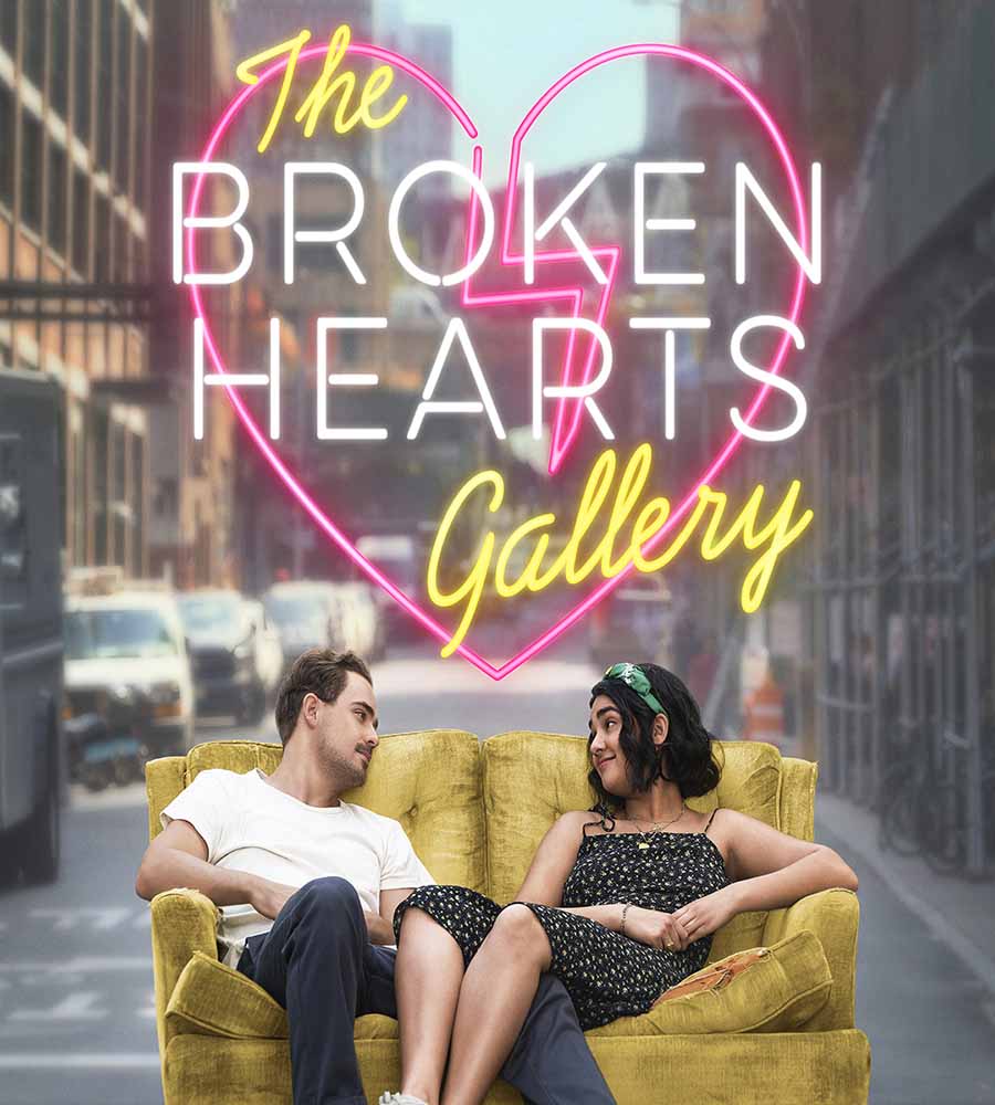 The image shows a promotional poster for "The Broken Hearts Gallery." In the foreground, a man and a woman sit casually on a mustard-yellow couch, gazing at each other with smiles. Behind them, the title "The Broken Hearts Gallery" is displayed in bold neon-style lettering, with a broken heart graphic surrounding the words. The background features a blurred urban street scene, adding to the romantic and urban vibe of the image.