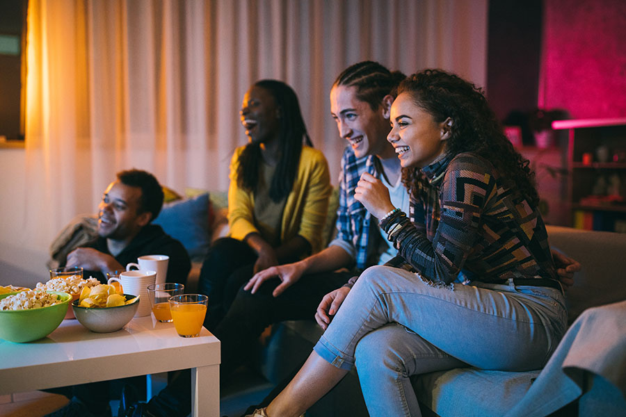 A diverse group of four friends, including people of different ethnicities, sit together on a couch, smiling and laughing while watching something on TV. A table in front of them holds snacks and drinks, suggesting a cozy and joyful evening.