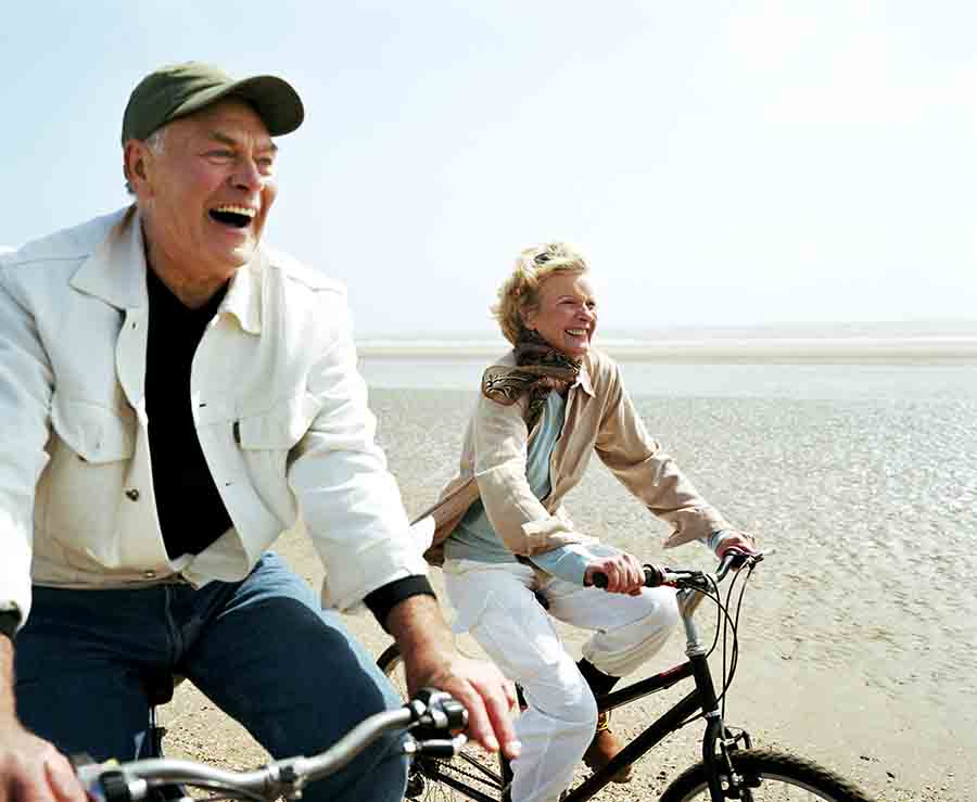 A joyful older couple rides bicycles along a sandy beach on a sunny day, laughing and enjoying the moment. The man wears a light jacket and cap, while the woman sports a scarf and beige coat.