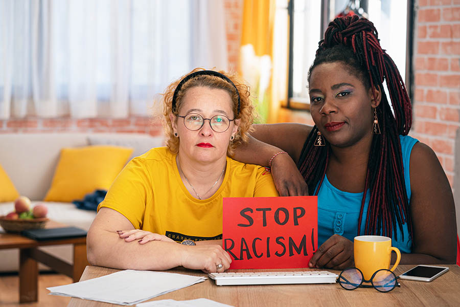 Two women, one white and one Black, sit together at a table holding a red sign that reads "STOP RACISM!" Both have serious and determined expressions, emphasizing unity and solidarity against racial injustice.