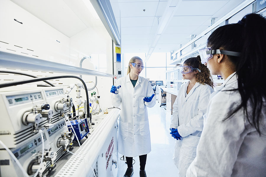 Three women scientists wearing white lab coats, safety goggles, and gloves are engaged in a discussion inside a bright, modern laboratory equipped with advanced scientific instruments.