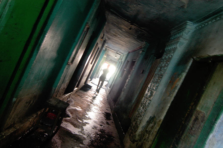 A dimly lit, narrow hallway with cracked and peeling green walls. A lone figure is seen walking towards a bright light at the end of the corridor, silhouetted against the glow. The scene suggests a deteriorating and confined space, evoking tension and unease.