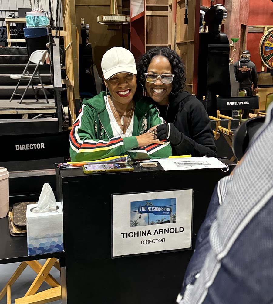 Zenay Arnold and Tichina Arnold smiling and posing together on the set of 'The Neighborhood,' seated behind a director’s table labeled with Tichina Arnold’s name and title, surrounded by set equipment and chairs.