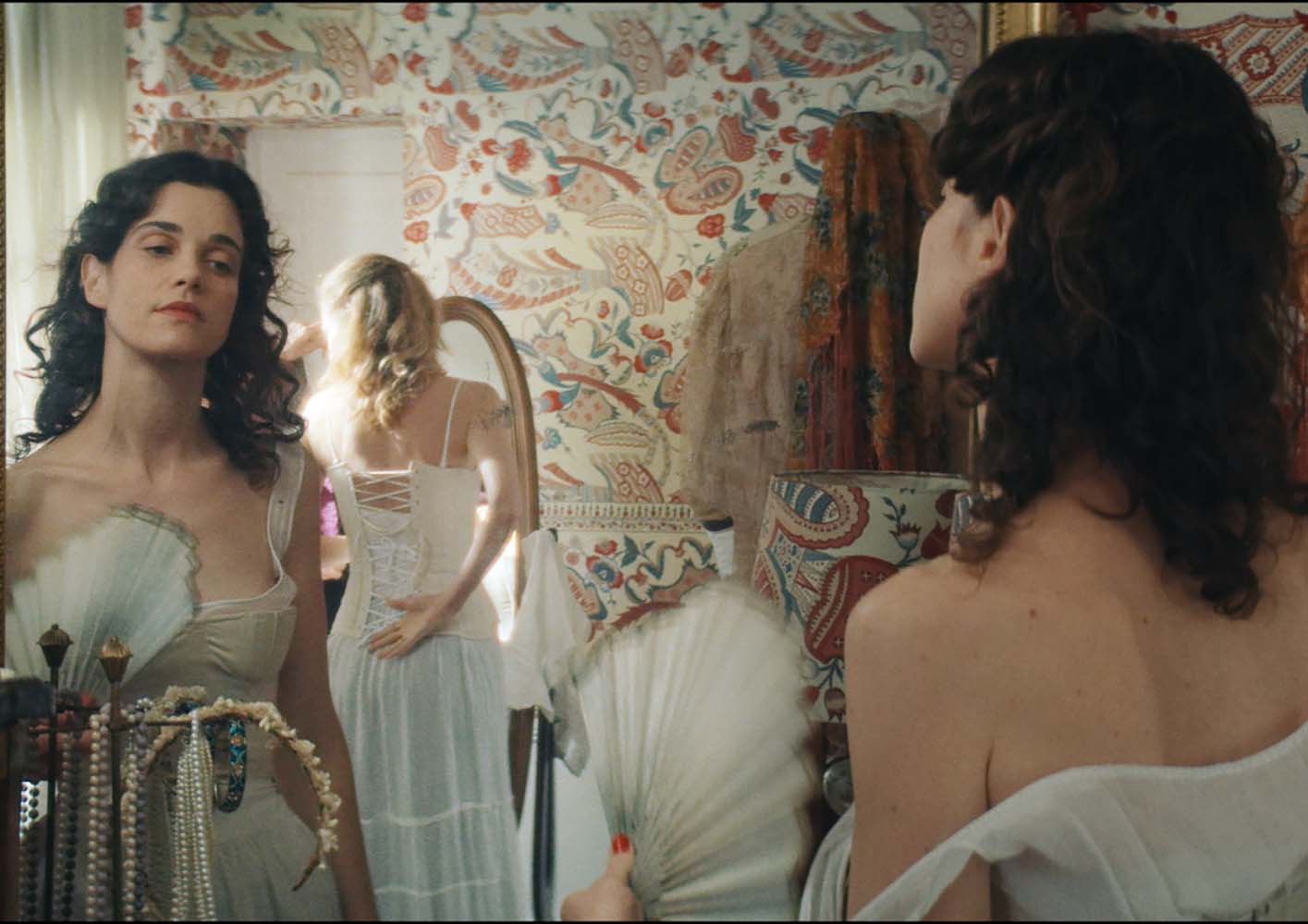 Two women dressed in vintage-style white corsets stand in front of ornate mirrors, adjusting their outfits. One woman looks confidently at her reflection while holding a delicate fan. The background features colorful, intricate paisley-patterned wallpaper and vintage décor.