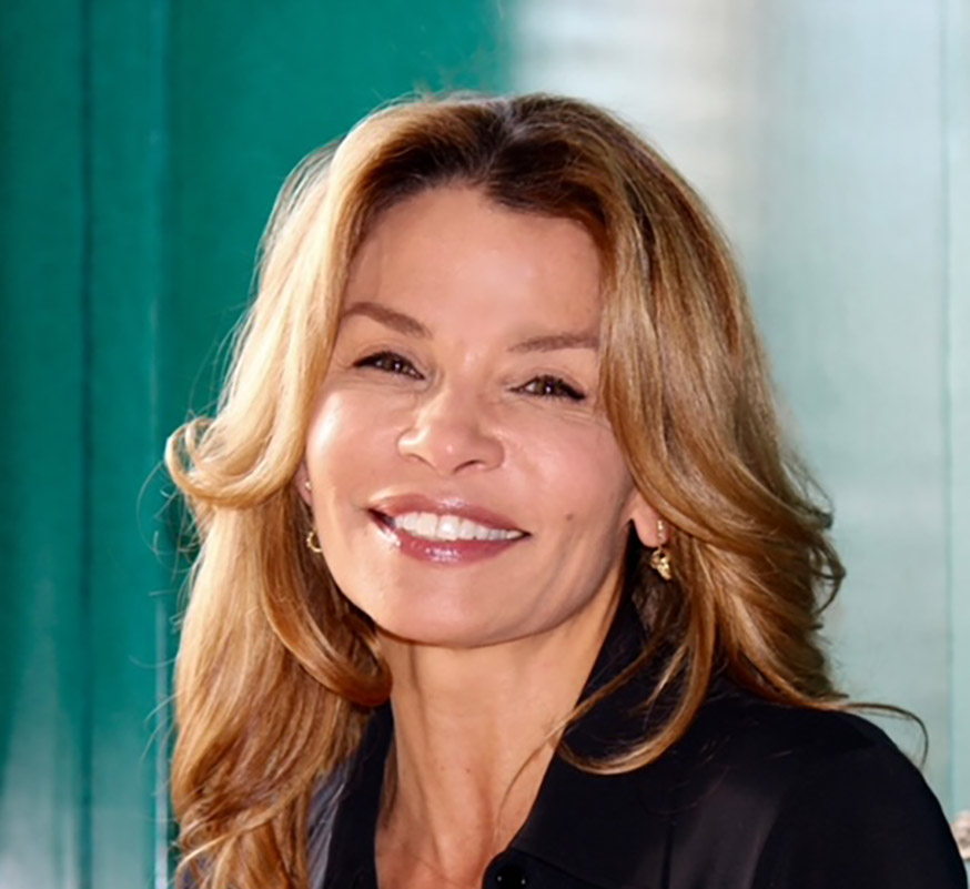 Jenny Lumet smiling, with long wavy light brown hair, wearing a black top and gold earrings, standing in front of a softly lit teal and glass background.