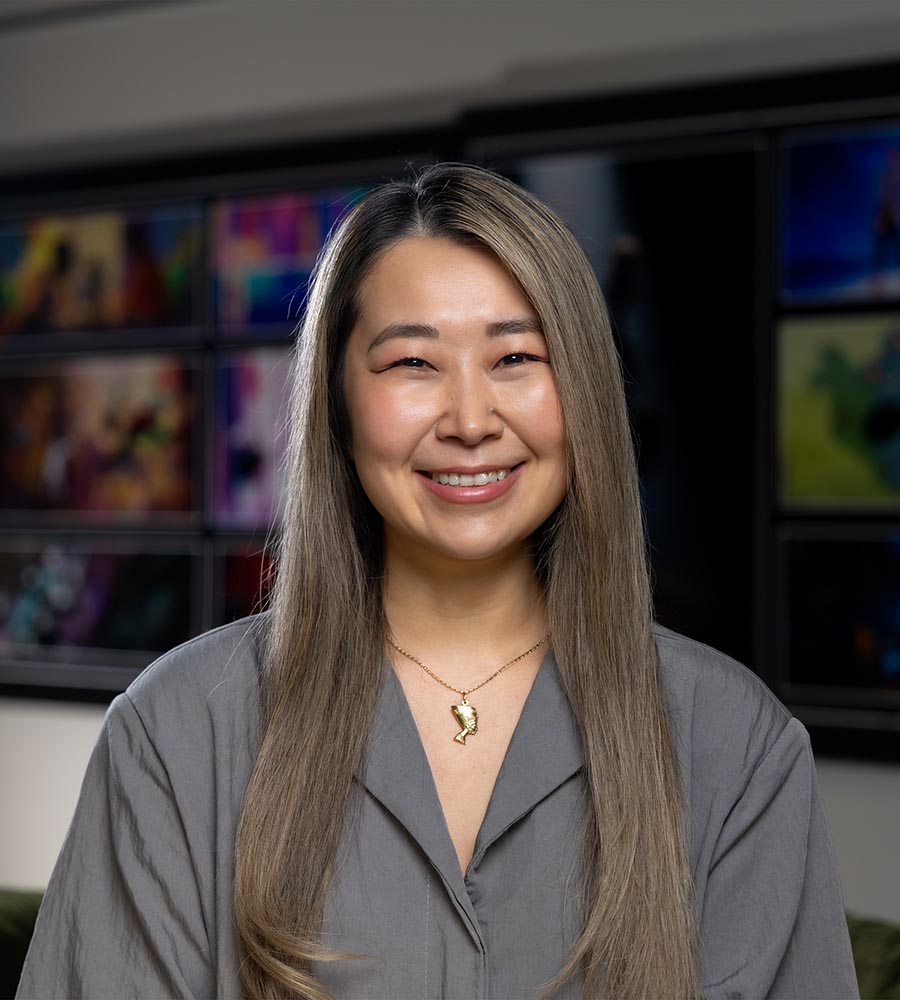 Maggie Kang, director at Sony Pictures Animation, smiling and wearing a gray blouse and gold pendant necklace, standing in front of a blurred background of colorful animation screens.