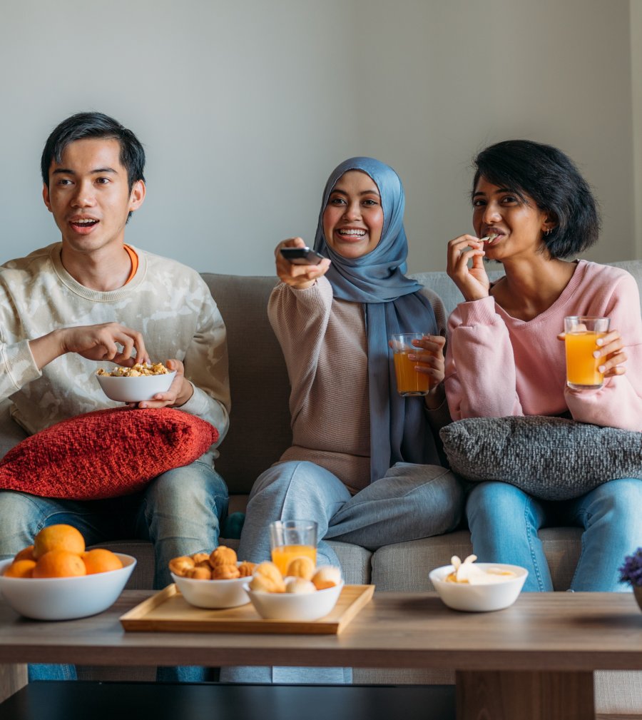 Image of an East Asian teen boy, a Muslim teen girl, and South Asian teen boy watching TV and sharing snacks