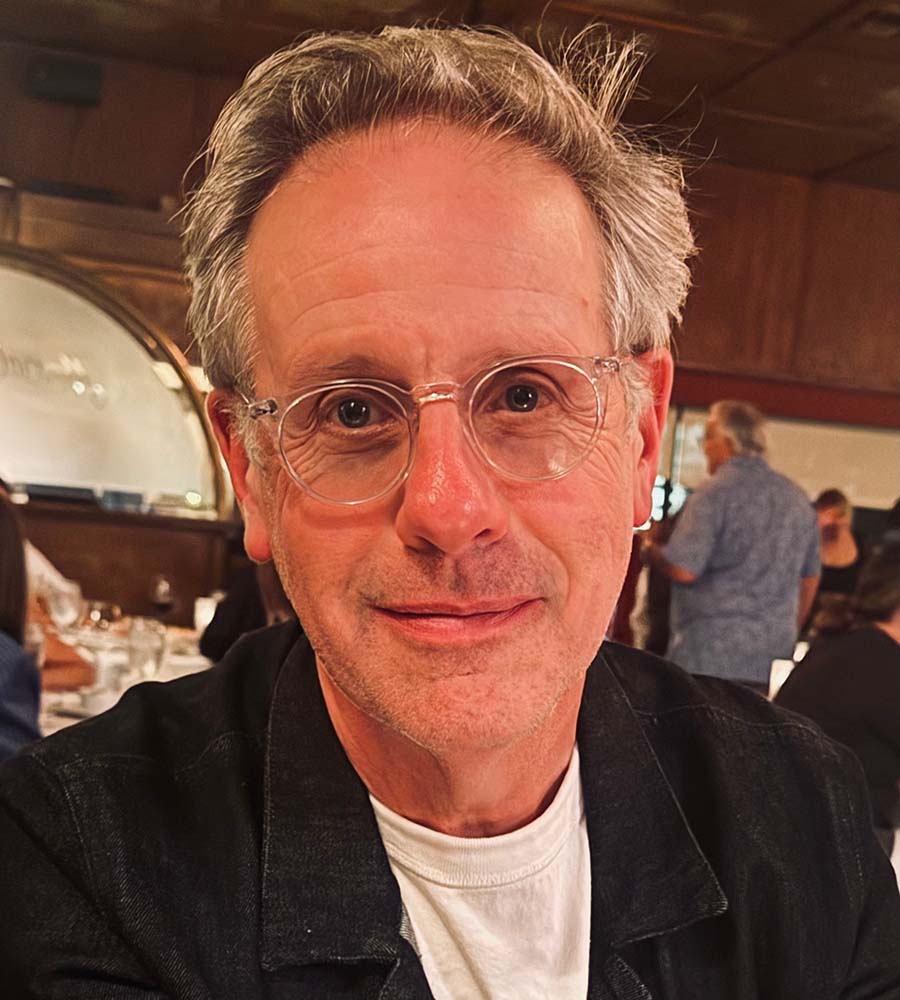 Headshot of film producer Peter Saraf wearing a dark denim blazer, smiling serenely with restaurant tables in background