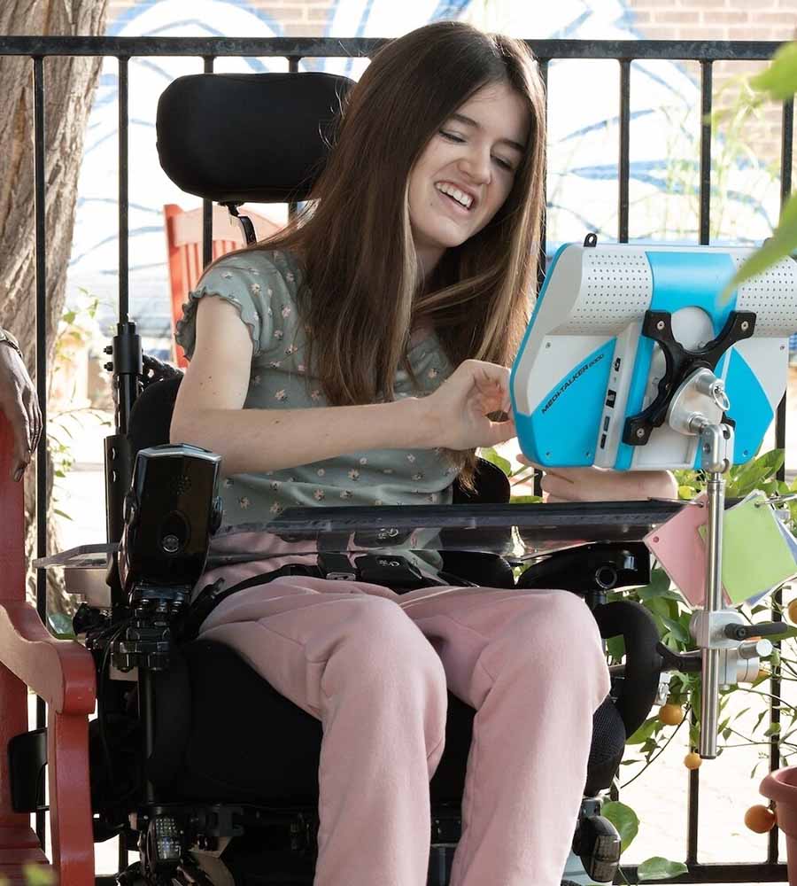 Teen girl with long brown hair sitting in a wheelchair, smiling while using a blue-and-white communication device, scene from Disney+ film Out of My Mind.