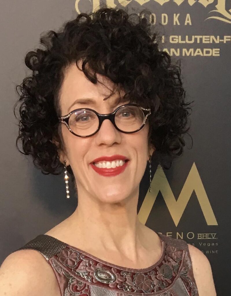Carin Greenberg smiling at a red carpet event, wearing glasses, dangling earrings, and a sleeveless dress with intricate maroon and silver embroidery, against a dark backdrop with gold and white text.