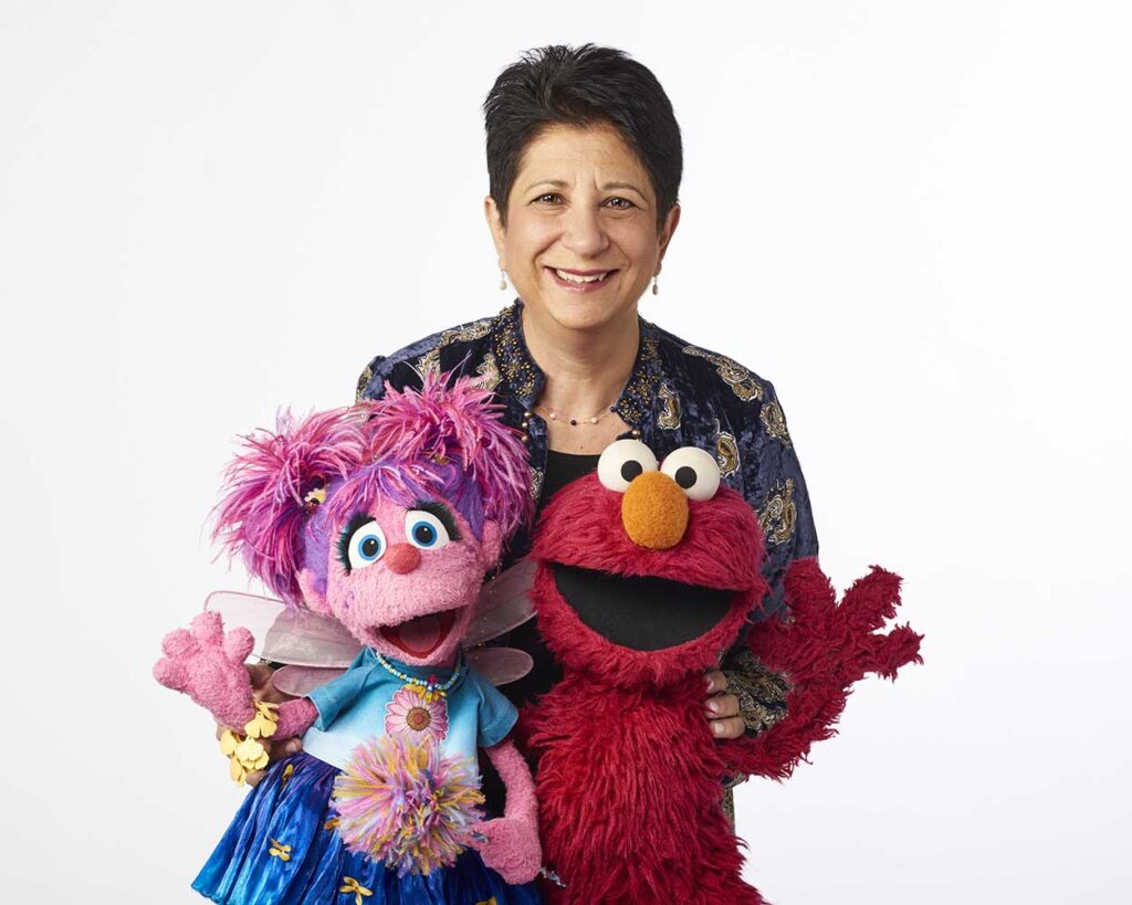 Rosemarie Truglio smiling while standing with Sesame Street characters Elmo and Abby Cadabby against a white background