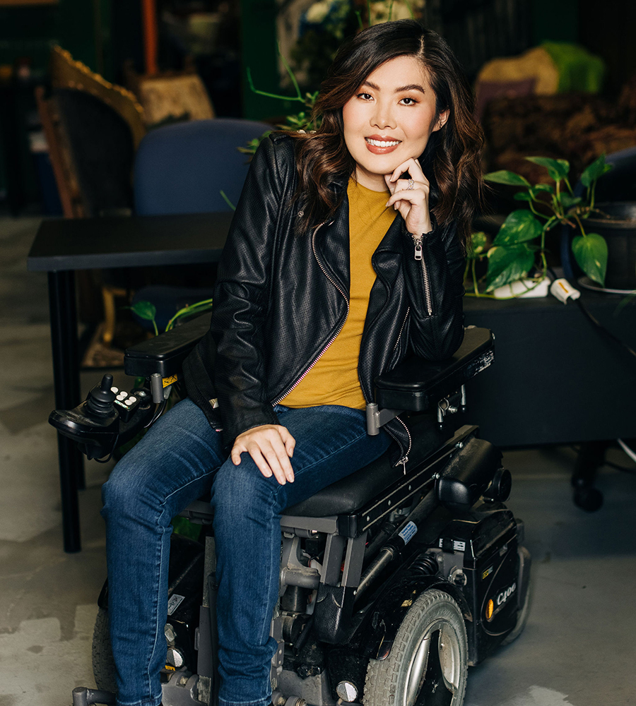 Kaitlyn Yang, a panelist from the Geena Davis Institute’s event on disability representation seated in a power wheelchair, smiling and wearing a black leather jacket and jeans in an indoor setting with plants.