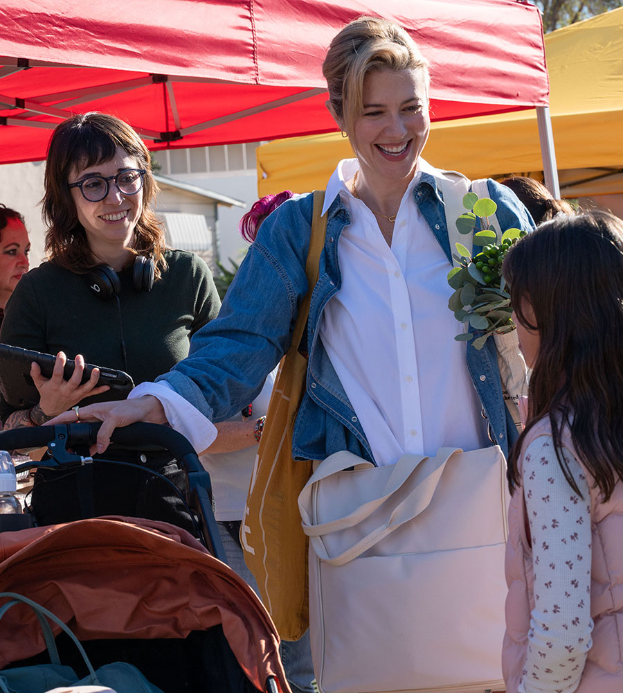 A professional behind-the-scenes photograph of director Michelle Garza Cervera on a film set. She is wearing a dark jacket and a black baseball cap, focused on a camera monitor while standing near a wooden cradle. In the background, the set is dimly lit, reflecting the atmospheric horror tone of the production.