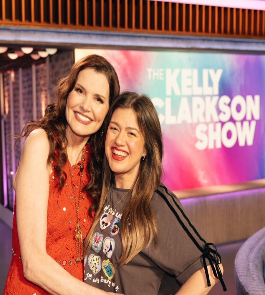 Geena Davis in a red lace dress smiling and hugging Kelly Clarkson on the vibrant set of The Kelly Clarkson Show.