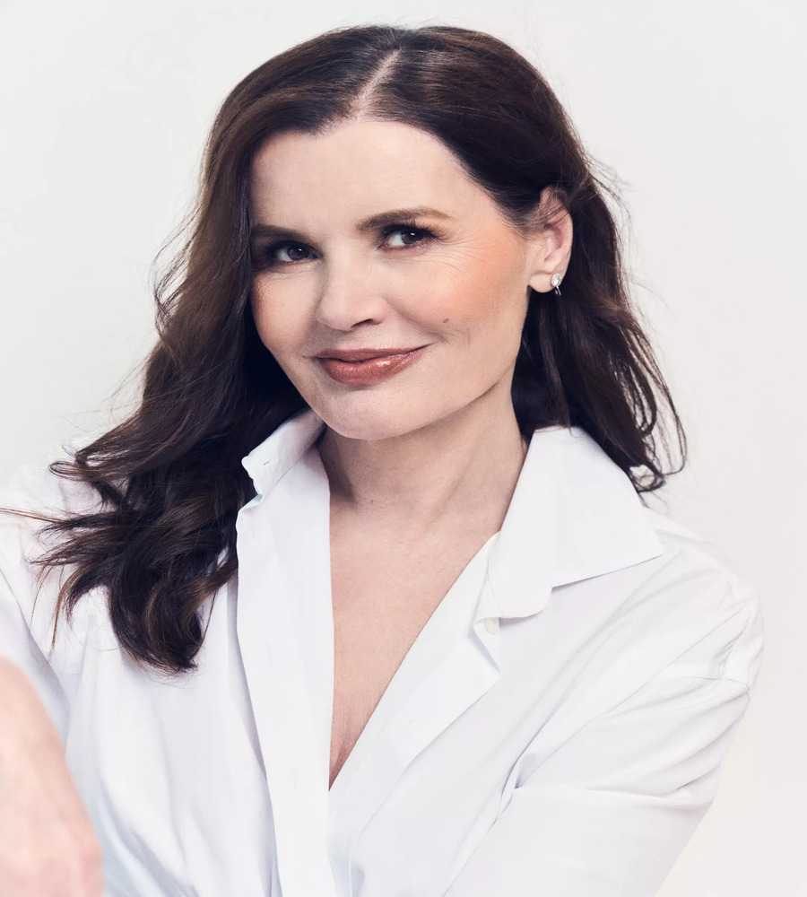 Studio portrait of Geena Davis looking directly at the camera with a subtle smile, wearing an open-collared white button-down shirt against a clean white backdrop.