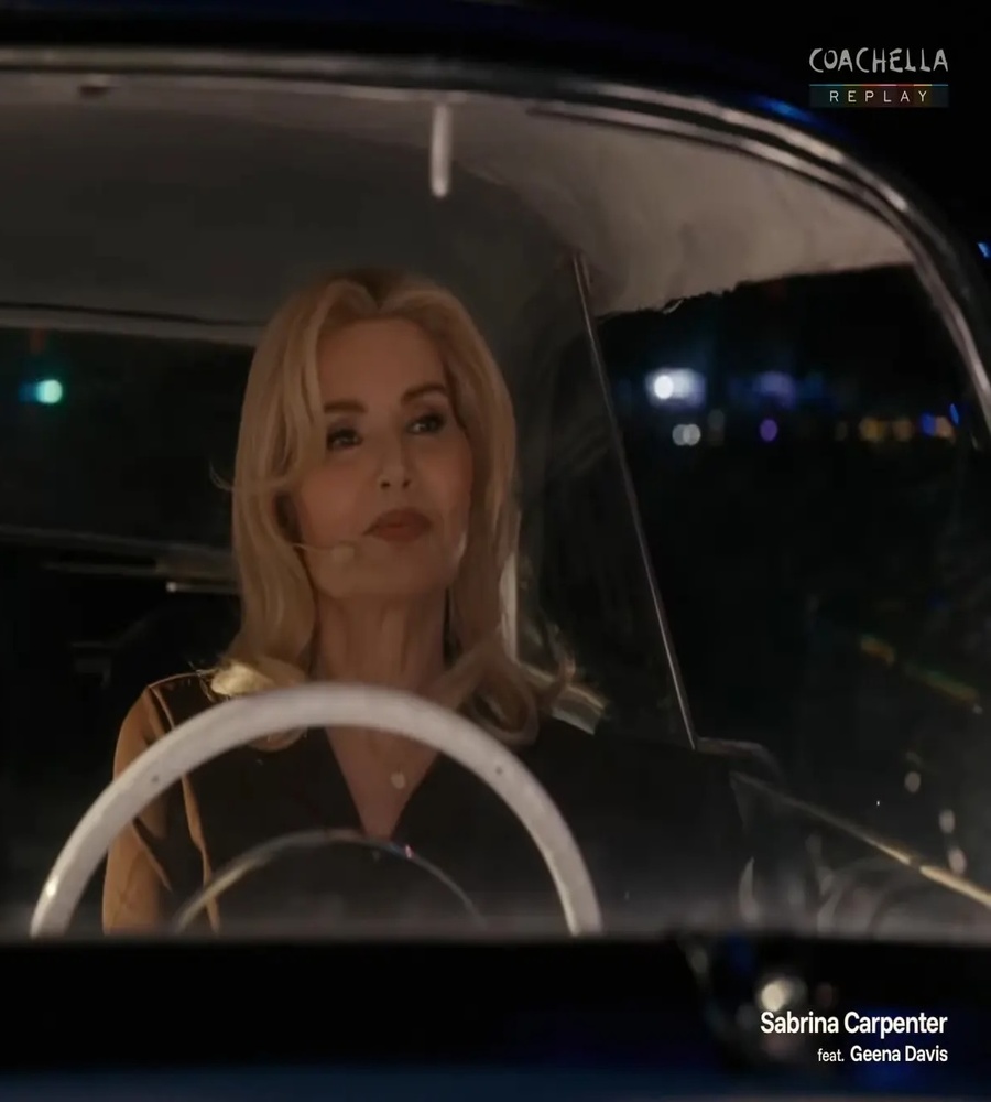 Screen capture from a Coachella broadcast showing Academy Award winner Geena Davis sitting behind the white steering wheel of a vintage convertible car at night, wearing a headset microphone as a guest performer during Sabrina Carpenter's musical set.