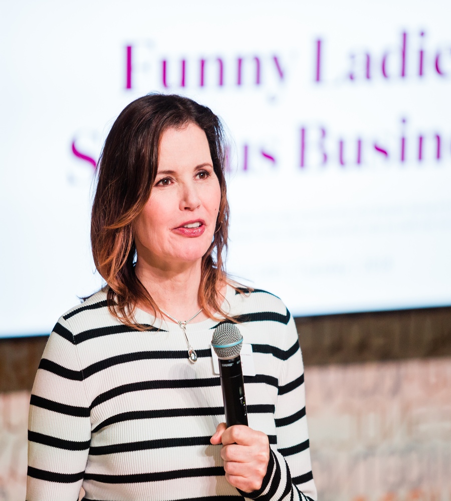 A medium close-up shot of actor and advocate Geena Davis speaking into a handheld microphone. She is wearing a white sweater with horizontal black stripes and a silver necklace. In the blurred background, a screen displays the event title "Funny Ladies: Serious Business" in purple text.