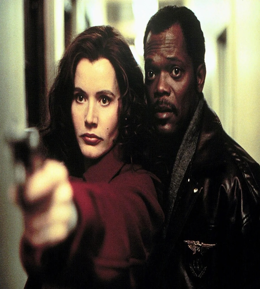 Actor Geena Davis stands in the foreground aiming a handgun forward, while a wide-eyed Samuel L. Jackson peers over her shoulder in a dark hallway.