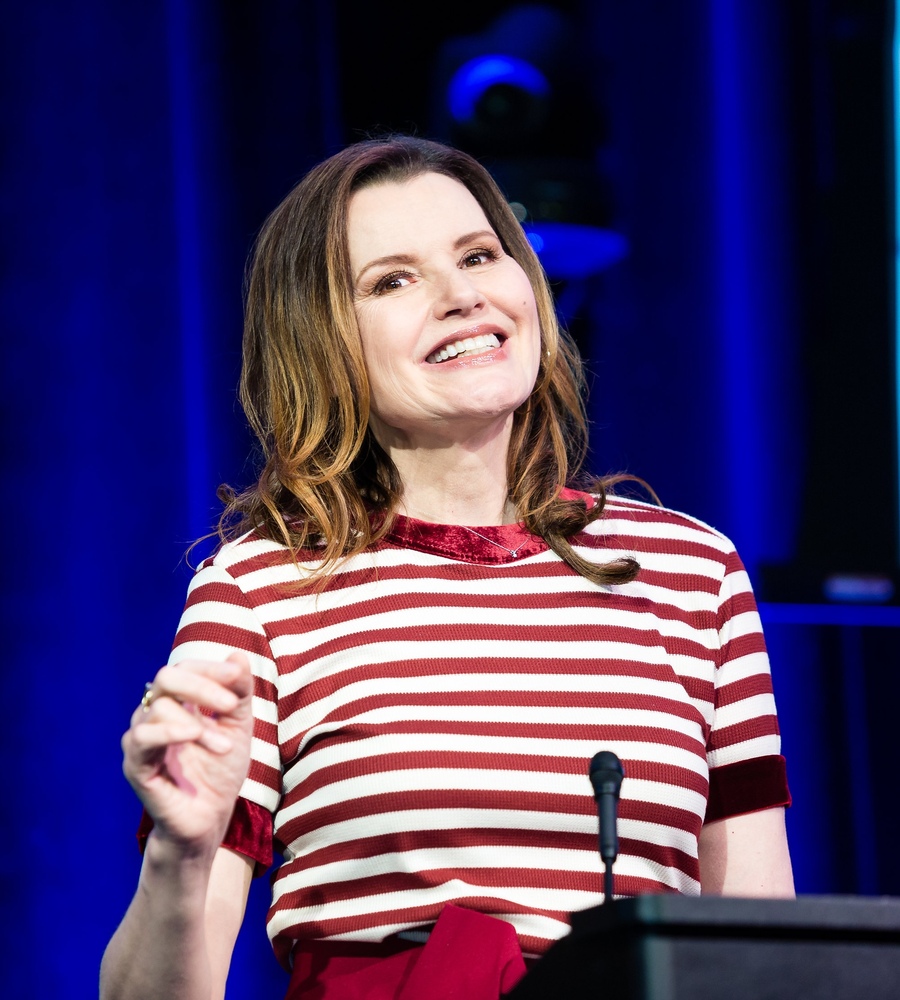 Geena Davis wearing a red and white striped top, smiling and speaking behind a podium at the Global Symposium on Gender in Media.