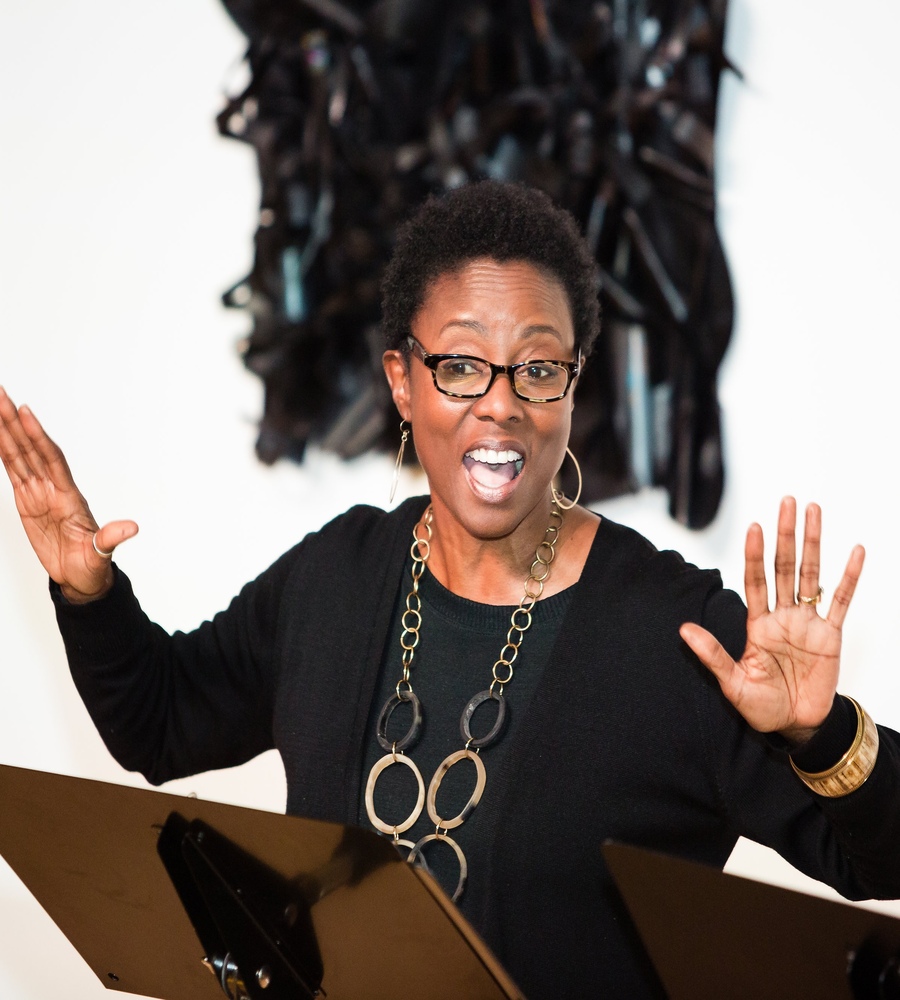 Joni Bovill gesturing expressive hands with an open mouth while performing a live dramatic reading behind a music stand at the See Jane Salon.